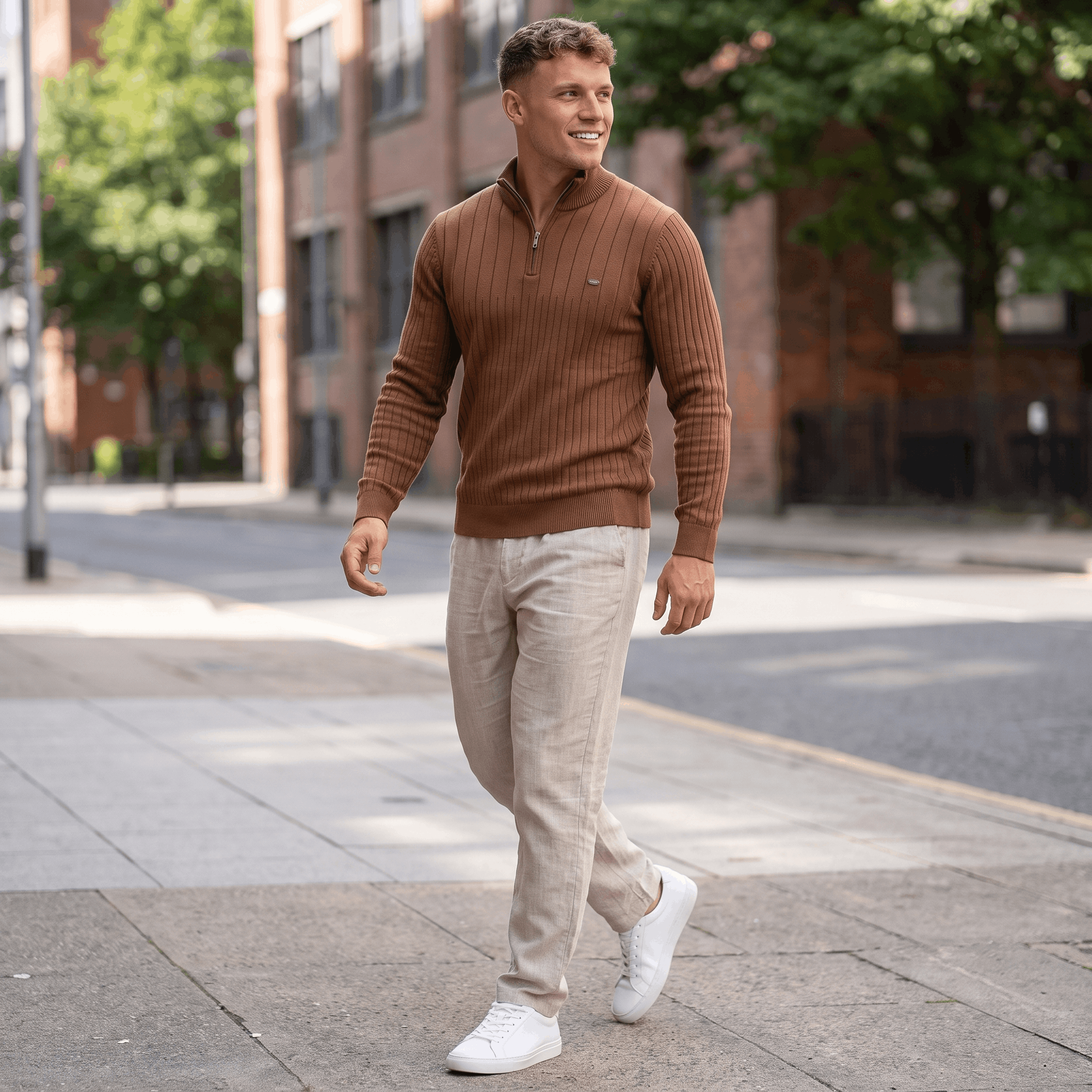 Man wearing a brown ribbed quarter-zip knit sweater with light-colored trousers walking along a city sidewalk.