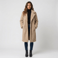 Woman wearing a beige long wool coat with wide lapels, front button closure, side pockets, and tailored silhouette over a black turtleneck, blue jeans, and black ankle boots.