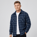 Man wearing a navy blue plaid overshirt jacket with a button-front closure, pointed collar, chest pockets, long sleeves, and relaxed fit over a white shirt and dark pants.