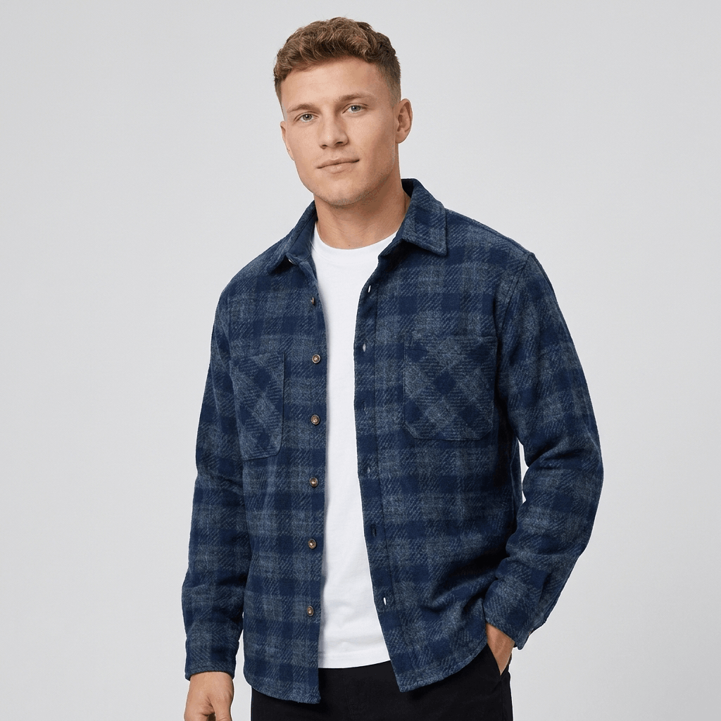 Man wearing a navy blue plaid overshirt jacket with a button-front closure, pointed collar, chest pockets, long sleeves, and relaxed fit over a white shirt and dark pants.